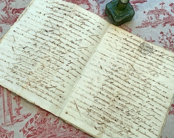 May include: An open antique book with handwritten script in brown ink. A small green glass inkwell with a white feather quill sits on the right side. The book and inkwell rest on a red and white patterned fabric.