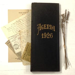 May include: A black rectangular 1926 agenda book with gold lettering on the cover, accompanied by a collection of vintage French paper documents, including a telegram and a small rectangular label, alongside a dried sprig of lavender.