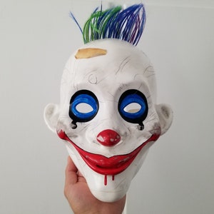 May include: A white plastic clown mask with blue eyes, a red smile, and a red nose. The mask has a green and purple mohawk and a small brown patch on the forehead.