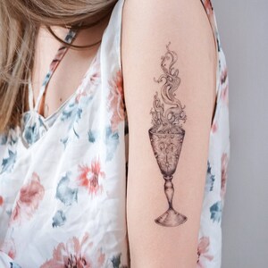 May include: A black and white tattoo of a goblet with smoke rising from it. The goblet is detailed with a pattern of stars and circles.
