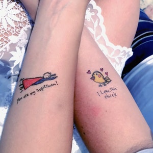 May include: Two arms with matching temporary tattoos. One arm features a cartoon Superman with the text "You are my superman!". The other arm has a yellow bird with hearts and the text "I love this chick".