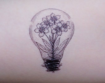 Puede incluir: Tatuaje en blanco y negro de una bombilla con flores en su interior. La bombilla está dibujada con un contorno detallado, y las flores tienen delicados pétalos y hojas. El tatuaje está en un brazo.