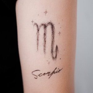 Op de afbeelding: Een zwarte inkt tattoo van het sterrenbeeld Schorpioen met een paar kleine sterren eromheen. Het woord "Schorpioen" staat in cursief onder het teken.