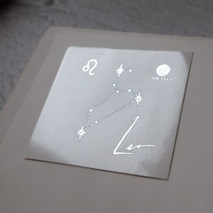 May include: A white sticker with a silver outline of the constellation Leo. The constellation is labeled with the astrological symbol for Leo and the word "Leo". The sticker also includes the text "Last Day" and a small circle with a "J" inside.