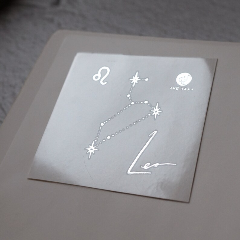 May include: A white sticker with a silver outline of the constellation Leo. The constellation is labeled with the astrological symbol for Leo and the word "Leo". The sticker also includes the text "Last Day" and a small circle with a "J" inside.
