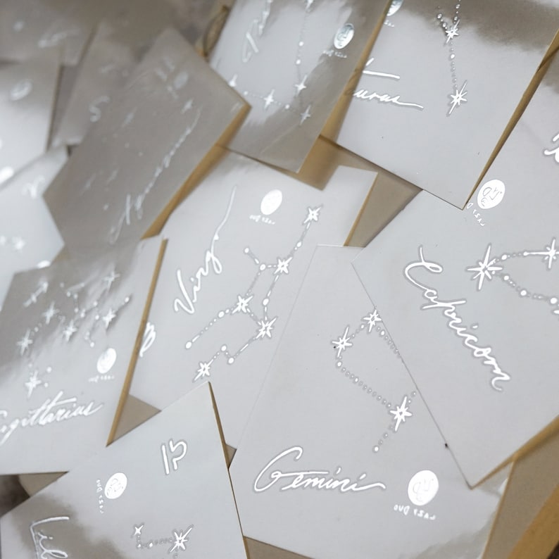 Op de afbeelding: Een set van twaalf witte kaarten met zilveren folielettering en sterrenbeeldontwerpen. Elke kaart heeft een ander sterrenbeeld, waaronder Tweelingen, Maagd, Boogschutter en Steenbok.