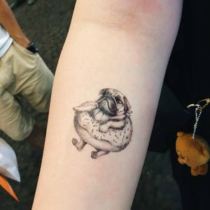 May include: A monochrome tattoo of a pug dog inside a donut. The tattoo is on an arm, showing the pug's face and paws. The donut has a dotted pattern, creating a playful design.