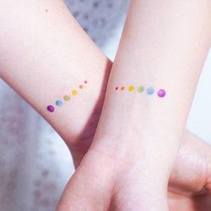 May include: Two wrists with small rainbow dot tattoos. The dots are in order of the rainbow colours, with purple, blue, green, yellow, orange, and red.