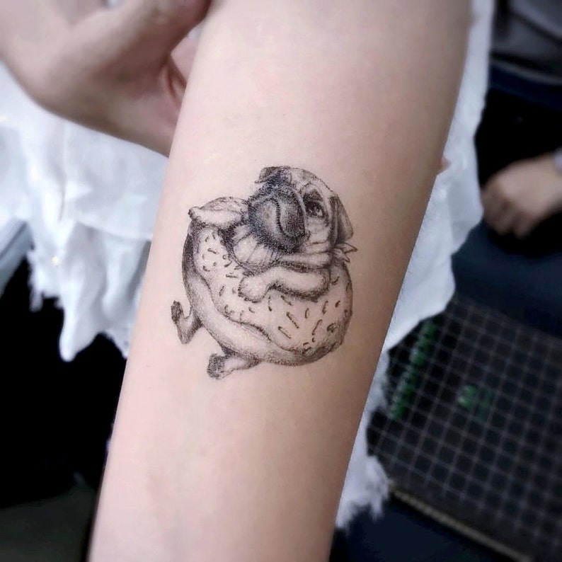 May include: A black and white tattoo of a pug dog inside a donut. The pug is lying down, with its paws and head visible. The donut has sprinkles. The tattoo is on a person's arm.