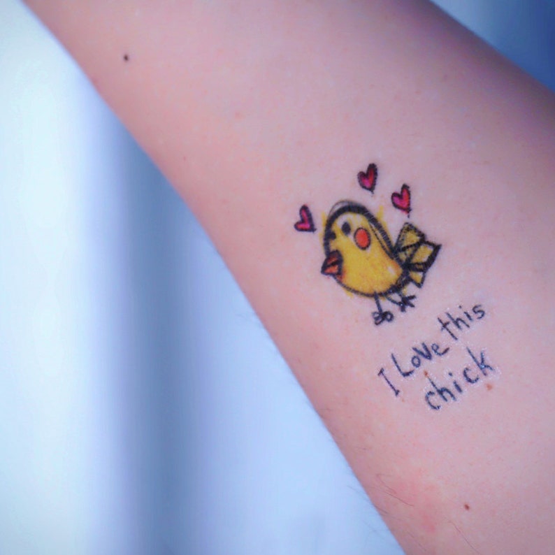 May include: A temporary tattoo featuring a yellow cartoon bird with a red beak and three red hearts. The text "I love this chick" is written below the bird in black ink.