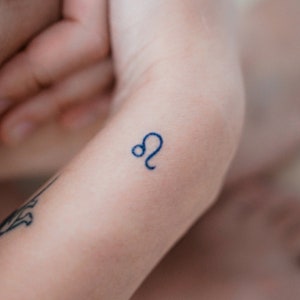 May include: A close-up of a small tattoo of the astrological symbol for Leo on a person's wrist.
