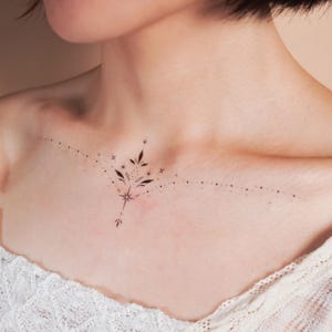 May include: A delicate tattoo design featuring a stylized floral branch with small stars and dots, placed on the collarbone.