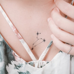 May include: A small, delicate tattoo of the constellation Leo on a person's chest. The tattoo is done in black ink and features a stylized outline of the constellation with small stars.