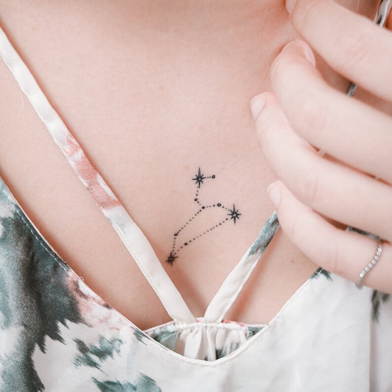 May include: A small, delicate tattoo of the constellation Leo on a person's chest. The tattoo is done in black ink and features a stylized outline of the constellation with small stars.