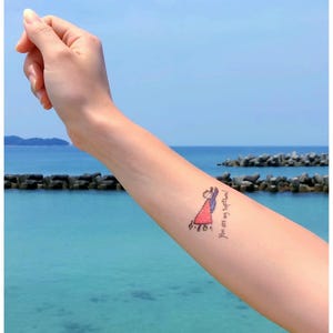 May include: A temporary tattoo on an arm showing a cartoon illustration of a person in a red dress and blue cape. The text "You are my Superman!" is written in cursive. The background shows the ocean and a clear blue sky.