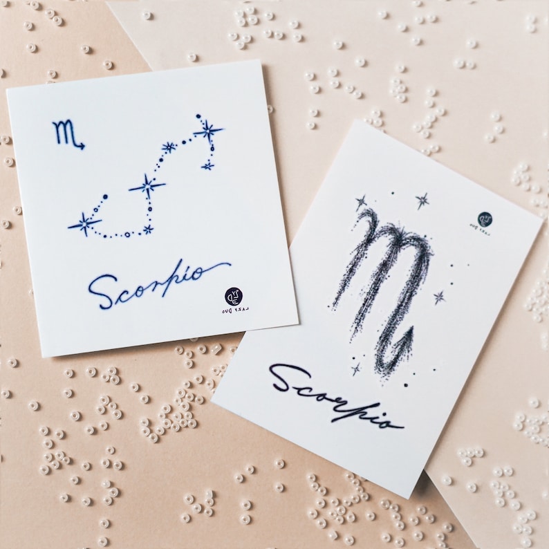 Op de afbeelding: Twee witte kaarten met blauwe inkt-illustraties van het sterrenbeeld Schorpioen. De eerste kaart toont een sterrenbeeld met de letter "m" en het woord "Schorpioen". De tweede kaart toont een meer abstracte illustratie van het Schorpioen-teken met het woord "Schorpioen".