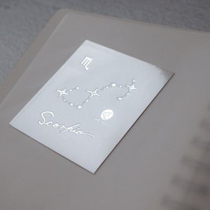 Op de afbeelding: Een zilveren sticker met een sterrenbeeld dat het sterrenbeeld Schorpioen voorstelt. De sticker bevat ook het symbool voor Schorpioen en het woord "Schorpioen" in cursief.