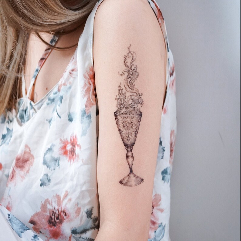 May include: A black and white tattoo of a goblet with smoke rising from it. The goblet is detailed with a pattern of stars and circles.