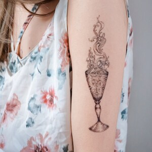 May include: A black and white tattoo of a goblet with flames rising from it on a person's arm.