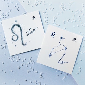 May include: Two white cards with blue ink illustrations of the Leo zodiac sign. One card shows a stylized Leo constellation with stars, the other shows a more abstract Leo design. Both cards have the word "Leo" written in cursive.