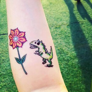 May include: A temporary tattoo showing a large flower with pink and yellow petals and a green stem. Beside the flower is a green dinosaur with a white skull design. The tattoo is on a person's arm.