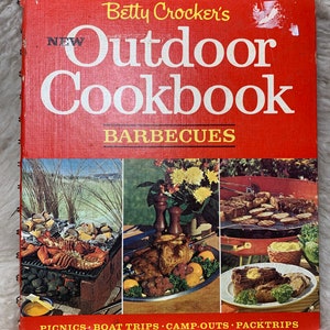 May include: A red cookbook with the title "Betty Crocker's New Outdoor Cookbook" and the subtitle "Barbecues". The cover features three images of food being cooked outdoors: lobster on a grill, a roasted chicken on a spit, and steak on a grill.