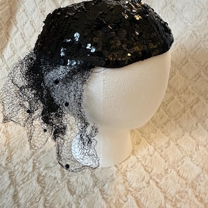 May include: A black sequined hat with a delicate black netting veil. The hat is displayed on a white mannequin head against a textured white background. The sequins on the hat shimmer, reflecting light.