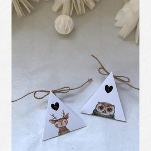 May include: Two white triangular gift boxes with illustrations of a deer and an owl, each with a black heart and tied with twine. The deer wears a sweater, and the owl wears a scarf. Decorative elements are in the background.