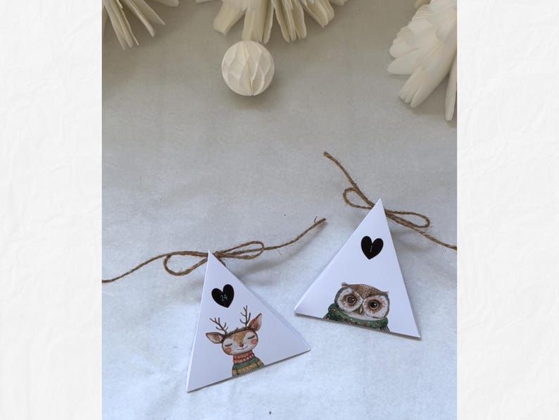 May include: Two white triangular gift boxes with illustrations of a deer and an owl, each with a black heart and tied with twine. The deer wears a sweater, and the owl wears a scarf. Decorative elements are in the background.