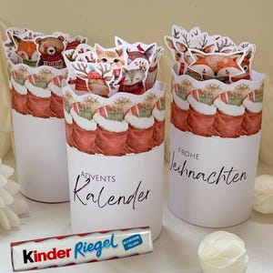 Könnte beinhalten: Drei weiße Adventskalender-Behälter mit Illustrationen von Weihnachtsstrümpfen und Waldtieren. Auf den Behältern stehen die Wörter "Advents Kalender" und "Frohe Weihnachten". Eine Kinder Riegel Schokoriegel befindet sich im Vordergrund.