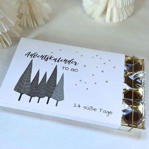 Peut inclure: Un calendrier de l'Avent blanc avec un motif de forêt et le texte "Adventskalender TO GO" et "24 süße Tage". La boîte contient plusieurs chocolats. Des étoiles dorées sont dispersées sur la boîte.