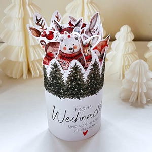 Puede incluir: Una tarjeta emergente blanca con temática navideña con un oso polar, un zorro y otras criaturas del bosque con suéteres rojos. La tarjeta tiene un diseño de bosque en la base y las palabras "Frohe Weihnachten" en escritura negra. Hay árboles de Navidad de papel blanco en el fondo.