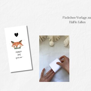 May include: A white card with a watercolor illustration of a sleeping fox with a green bow tie and the text "...nur noch schlafen!" A black heart is above the fox. The image also shows hands folding a white paper.