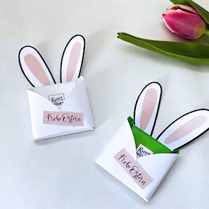 Könnte beinhalten: Zwei weiße Oster-Geschenktüten aus Papier mit Hasenohren. Jede Tüte hat ein rosa Etikett mit den Worten "Frohe Ostern". Eine rosa Tulpe und grüne Blätter sind im Hintergrund. Die Tüten enthalten Schokolade.
