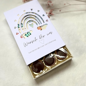 May include: A white gift box with a rainbow cutout design, filled with three chocolate candies. The rainbow design incorporates currency notes. The box has text that reads "Wunsch Dir was" and "Von Herzen alles Gute zum Geburtstag."
