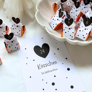 Può includere: Un calendario dell'Avvento con piccoli regali confezionati. Ogni regalo è bianco con pois neri e un'etichetta a forma di cuore nero con un numero. È presente anche una carta con un cuore nero e il numero 24.