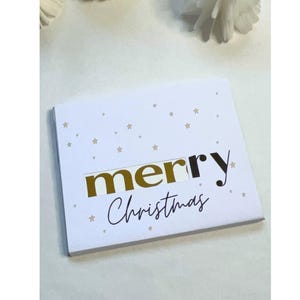 Puede incluir: Tarjeta navideña blanca con estrellas doradas y las palabras "merry Christmas" en oro y negro. La palabra "merry" está en un diseño recortado. Una tarjeta de felicitación festiva.