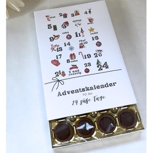 Könnte beinhalten: Adventskalender-Box mit weißem Deckel mit festlichen Illustrationen und den Zahlen 1-24. Die Box ist geöffnet und zeigt vier Pralinen. Auf dem Deckel steht "Adventskalender" und "24 süße Tage".