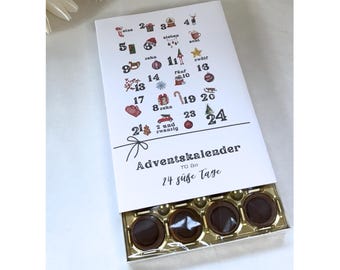 May include: Advent calendar box with a white lid featuring festive illustrations and numbers 1-24. The box is open, revealing four chocolate candies. The text "Adventskalender" and "24 süße Tage" are printed on the lid.
