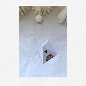May include: A white, geometric gift box with a black heart and an owl illustration. The box is folded into a pyramid shape and has small holes near the top. Decorative paper flowers and a white ball are in the background.