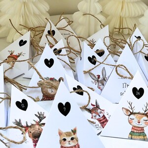 May include: Advent calendar with white triangular boxes, each adorned with a black heart and a number. The boxes feature illustrations of woodland animals, including a deer, cat, and bear, with twine closures. White paper Christmas trees are in the background.