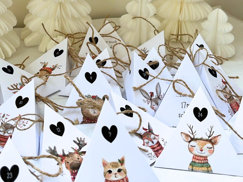 May include: Advent calendar with white triangular boxes, each adorned with a black heart and a number. The boxes feature illustrations of woodland animals, including a deer, cat, and bear, with twine closures. White paper Christmas trees are in the background.