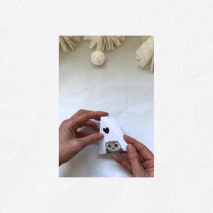 May include: A white paper craft shaped like a house, held by hands. The house has a black heart and an owl illustration. Decorative elements are in the background.