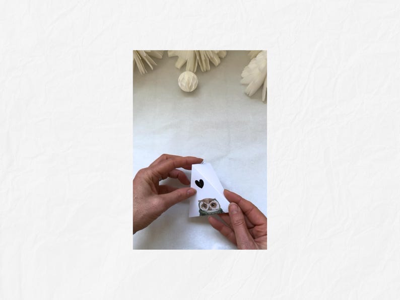 May include: A white paper craft shaped like a house, held by hands. The house has a black heart and an owl illustration. Decorative elements are in the background.