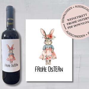 Op de afbeelding: Afdrukbaar wijnlabel met een aquarel-illustratie van een konijn in een roze jurk en een bloemenkrans. De tekst "Frohe Ostern" staat in het zwart onder het konijn.