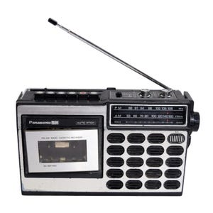 May include: A black and silver Panasonic LTD radio cassette recorder featuring a retractable antenna, a cassette deck with auto stop, and a speaker grille with twenty circular openings. The device includes AM and FM frequency tuning scales.