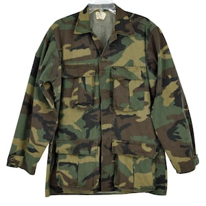 May include: A long-sleeved button-up shirt featuring a woodland camouflage pattern in shades of olive green, brown, and black. The shirt includes two chest pockets with flaps and two larger lower pockets with flaps, along with buttoned cuffs.