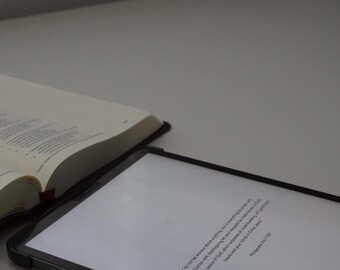 May include: An open book and a tablet with text are on a white surface. The book has yellowed pages and a dark brown cover. The tablet displays text from Philippians 4:6-7. The image has a soft, neutral color palette.