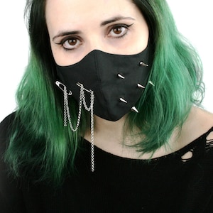 May include: Black face mask featuring silver spikes and chain detailing. The mask has a punk rock style, with safety pins and chains suspended from the lower edge. The mask is worn by a person with green hair.