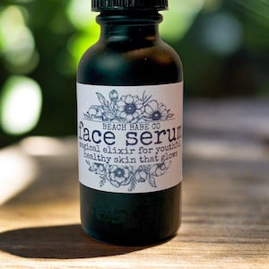 Puede incluir: Una peque&ntilde;a botella de vidrio verde oscuro con una tapa negra. La botella tiene una etiqueta blanca con texto negro que dice "Beach Babe Co. face serum magical elixir for youthful healthy skin that glows". La etiqueta est&aacute; decorada con un dise&ntilde;o floral.